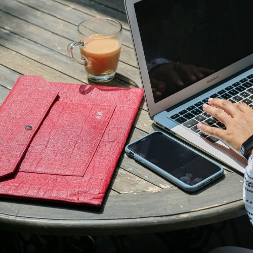 Mulberry coloured laptop sleeve on a wooden table next to a person drinking a coffee working on their laptop